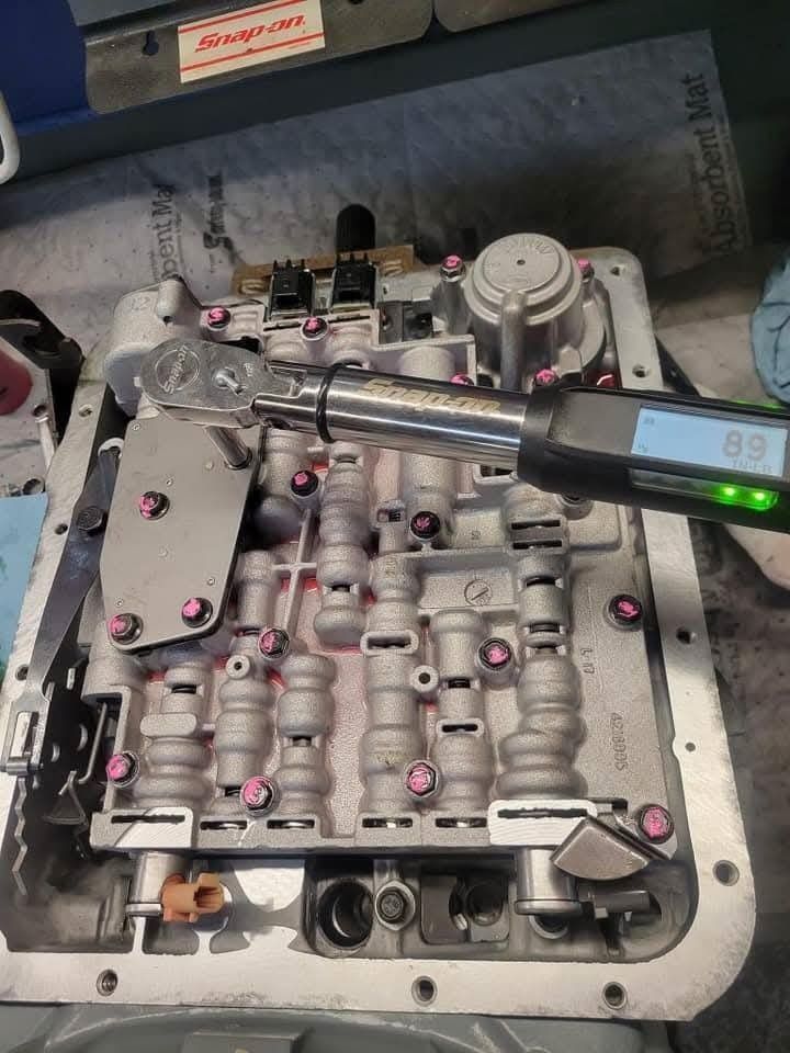 Mechanic using a torque wrench on a transmission valve body, pink bolts visible.
