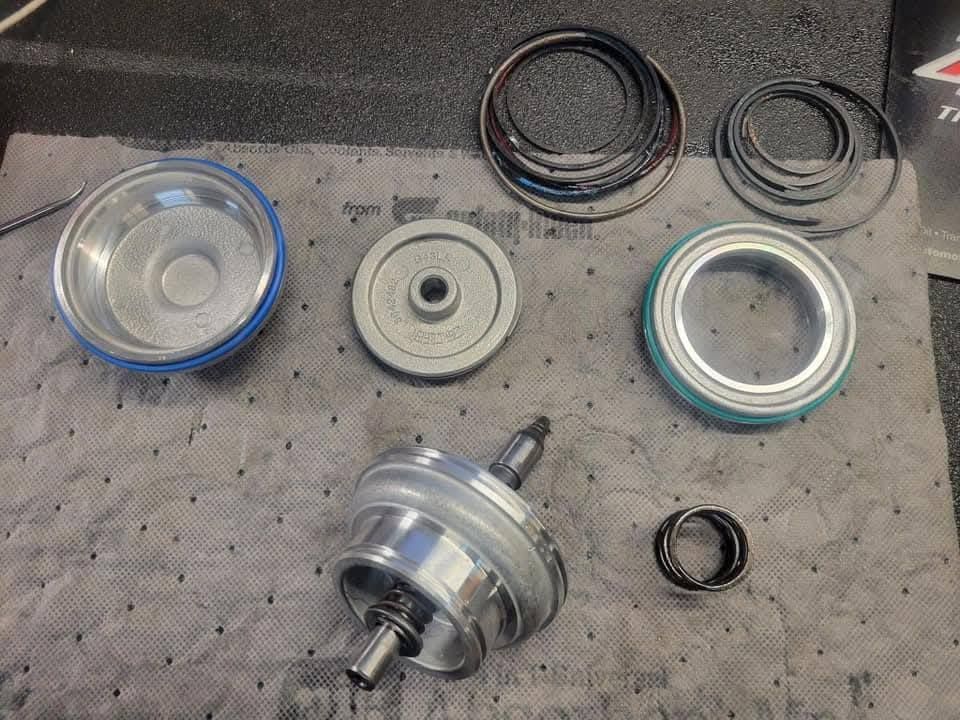 Disassembled vehicle part components laid on a gray absorbent mat. Includes metal and rubber rings, a bearing, and a metal cylinder.