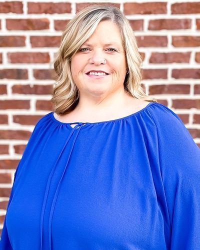 A woman in a blue shirt is smiling in front of a brick wall.