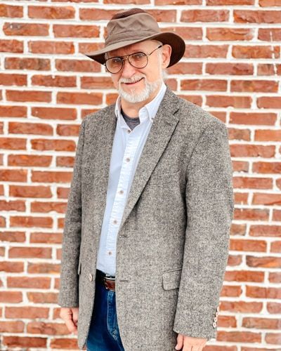 A man wearing a hat and glasses is standing in front of a brick wall.