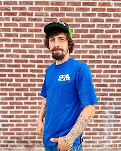 A man wearing a blue t-shirt and a hat is standing in front of a brick wall.