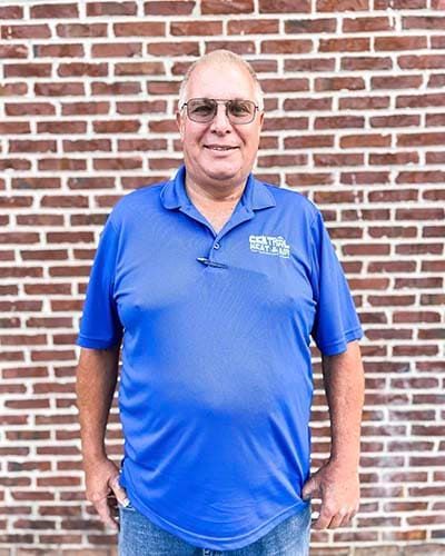 A man in a blue shirt and sunglasses is standing in front of a brick wall.