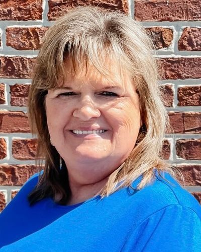 A woman in a blue shirt is smiling in front of a brick wall.