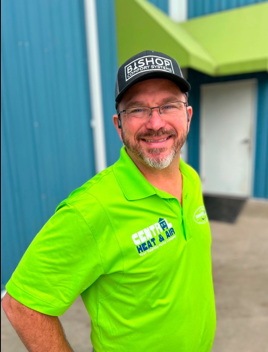A man in a blue shirt and green hat is standing in front of a brick wall with his arms crossed.