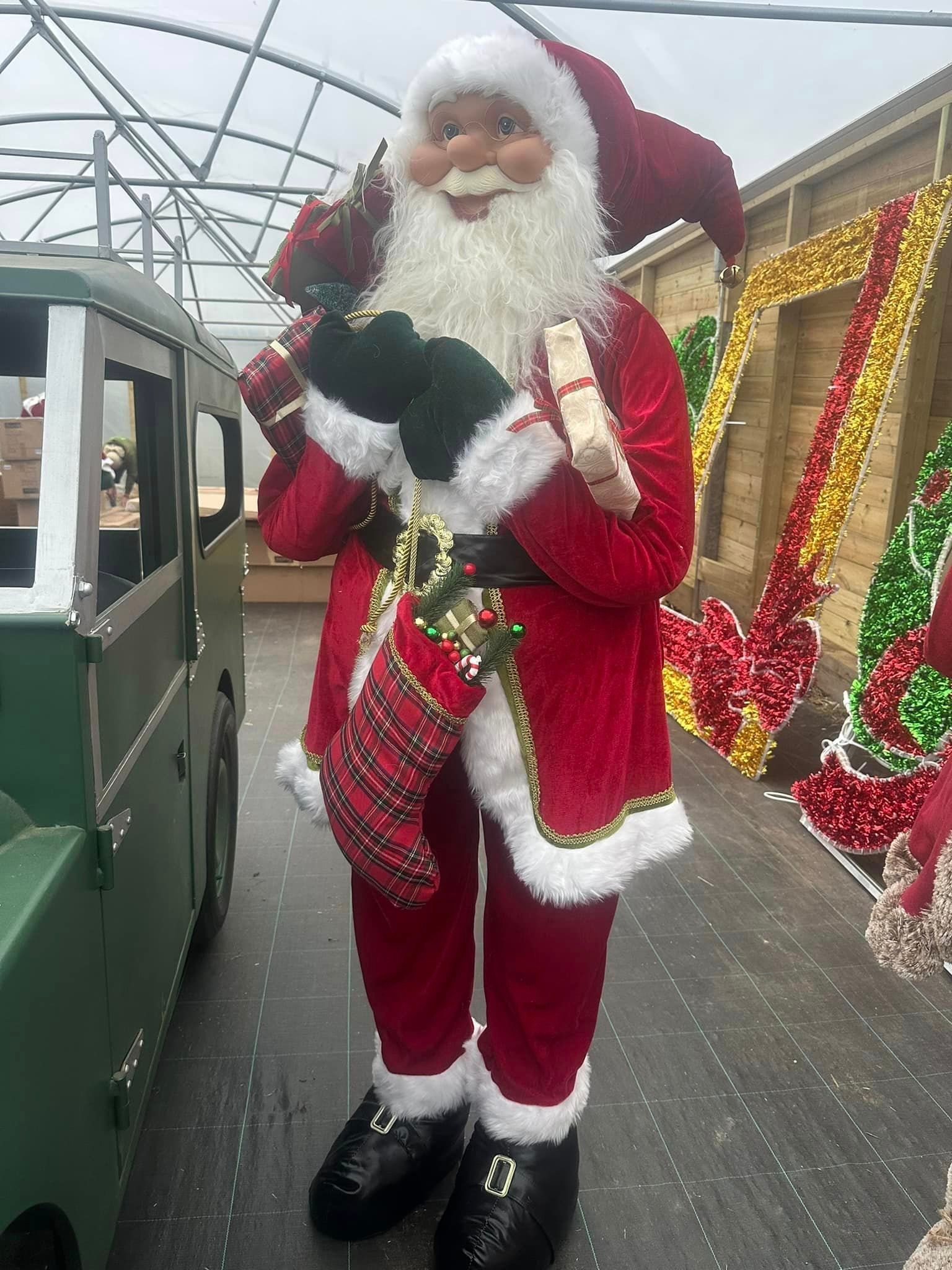 Santa Claus statue, red suit with gifts, standing near a green truck, Christmas decorations.