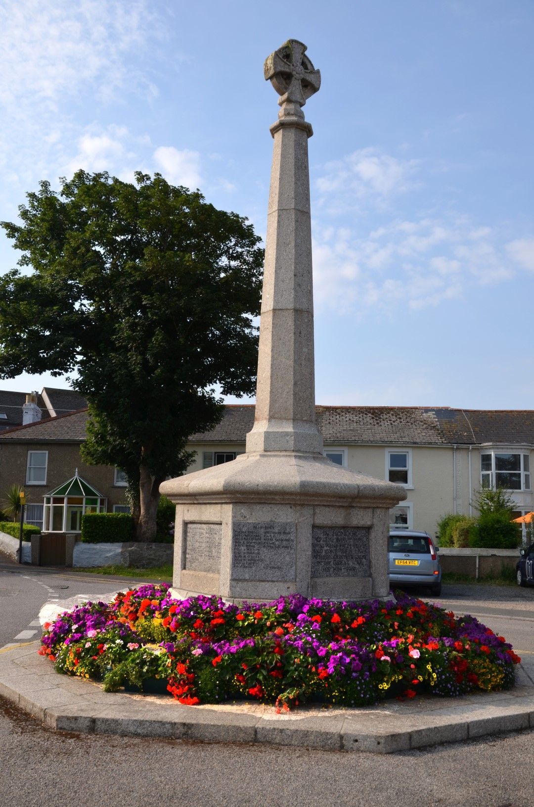 War memorial with cross on top, surrounded by a flowerbed, in front of buildings.