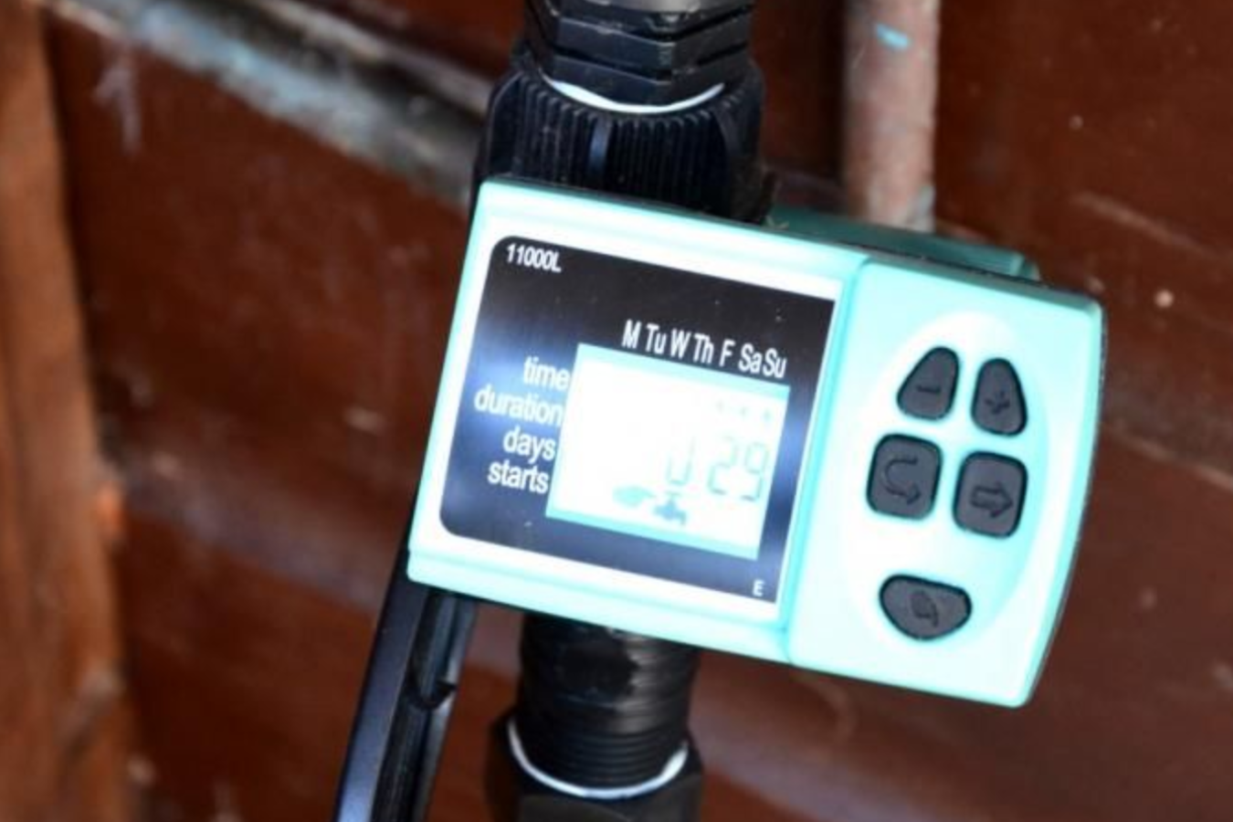 A turquoise digital water timer attached to a pipe against a brown wooden wall.