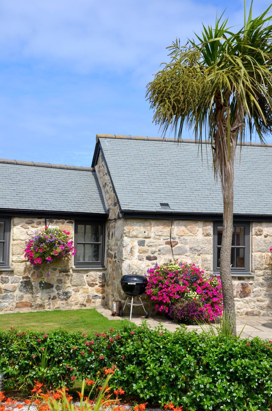 Stone cottage with hanging basket, barbecue, and palm tree under blue sky.