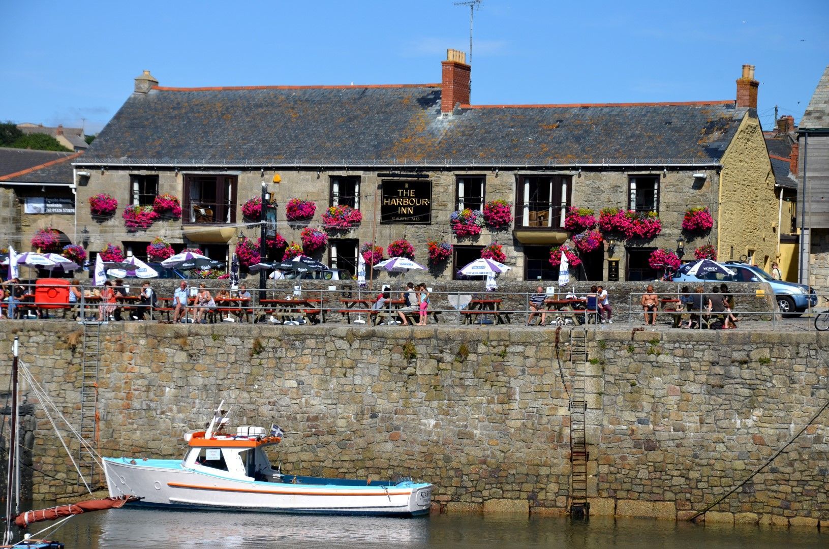 A stone building with red flower boxes, outdoor seating, and a boat in a harbor.