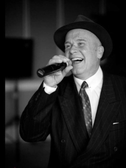 Man singing into a microphone, wearing a suit and hat, smiling broadly.