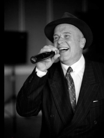 Man singing into a microphone, wearing a suit and hat, smiling broadly.