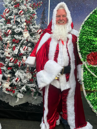 Man in Santa suit smiles, stands by snowy Christmas tree and large green ornament.