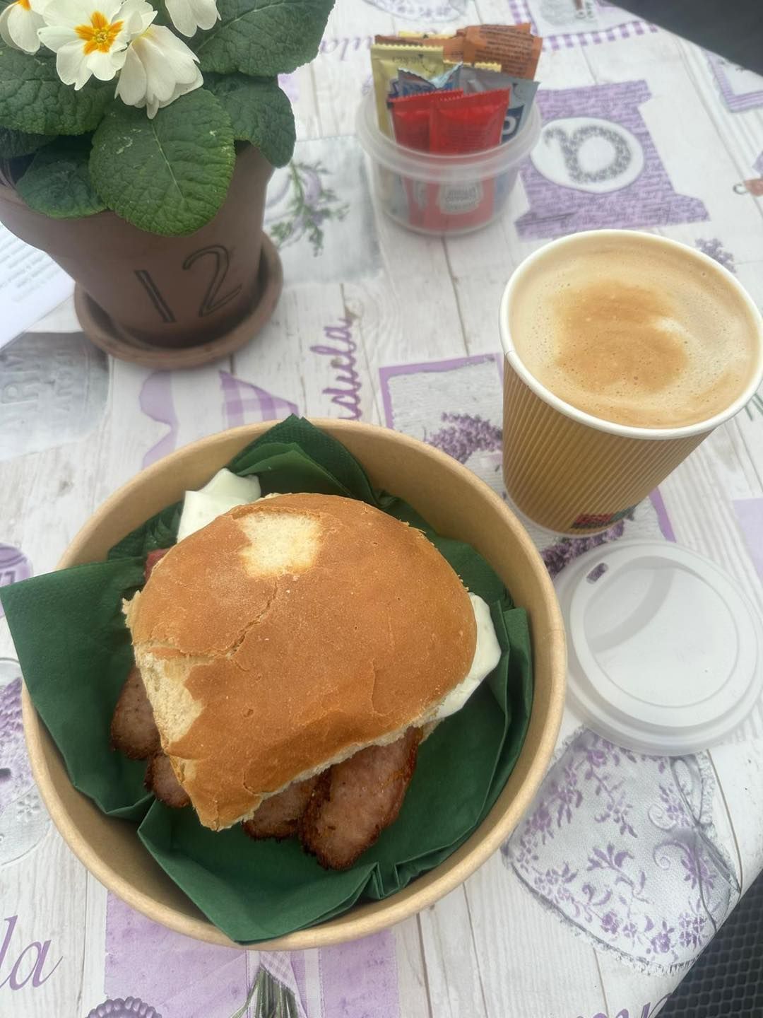 Breakfast: Sausage and egg sandwich in a bowl, coffee, condiments, and a flowerpot on a lavender tablecloth.