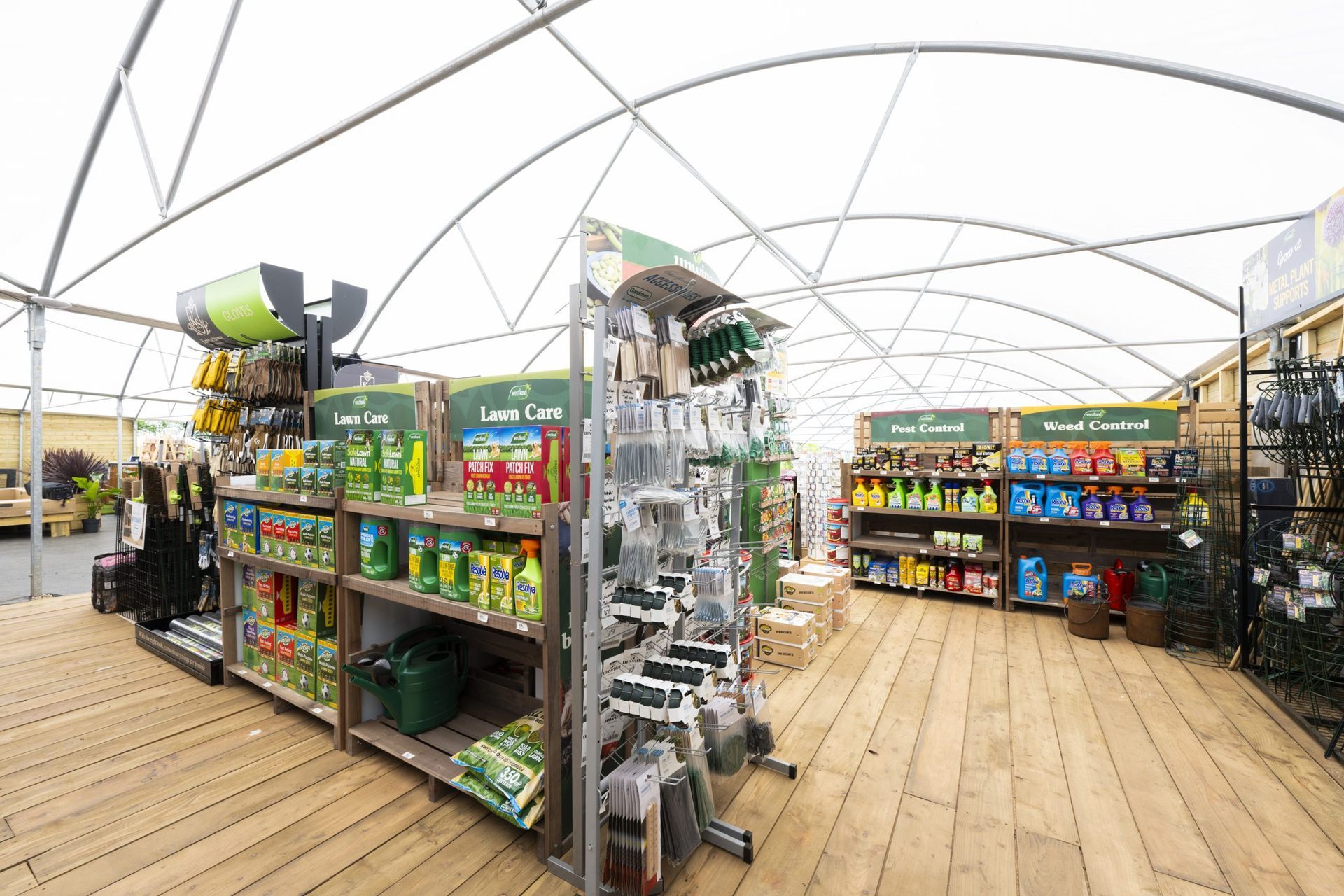 Interior of a garden center: shelves with gardening supplies, wooden floor, white arched roof.