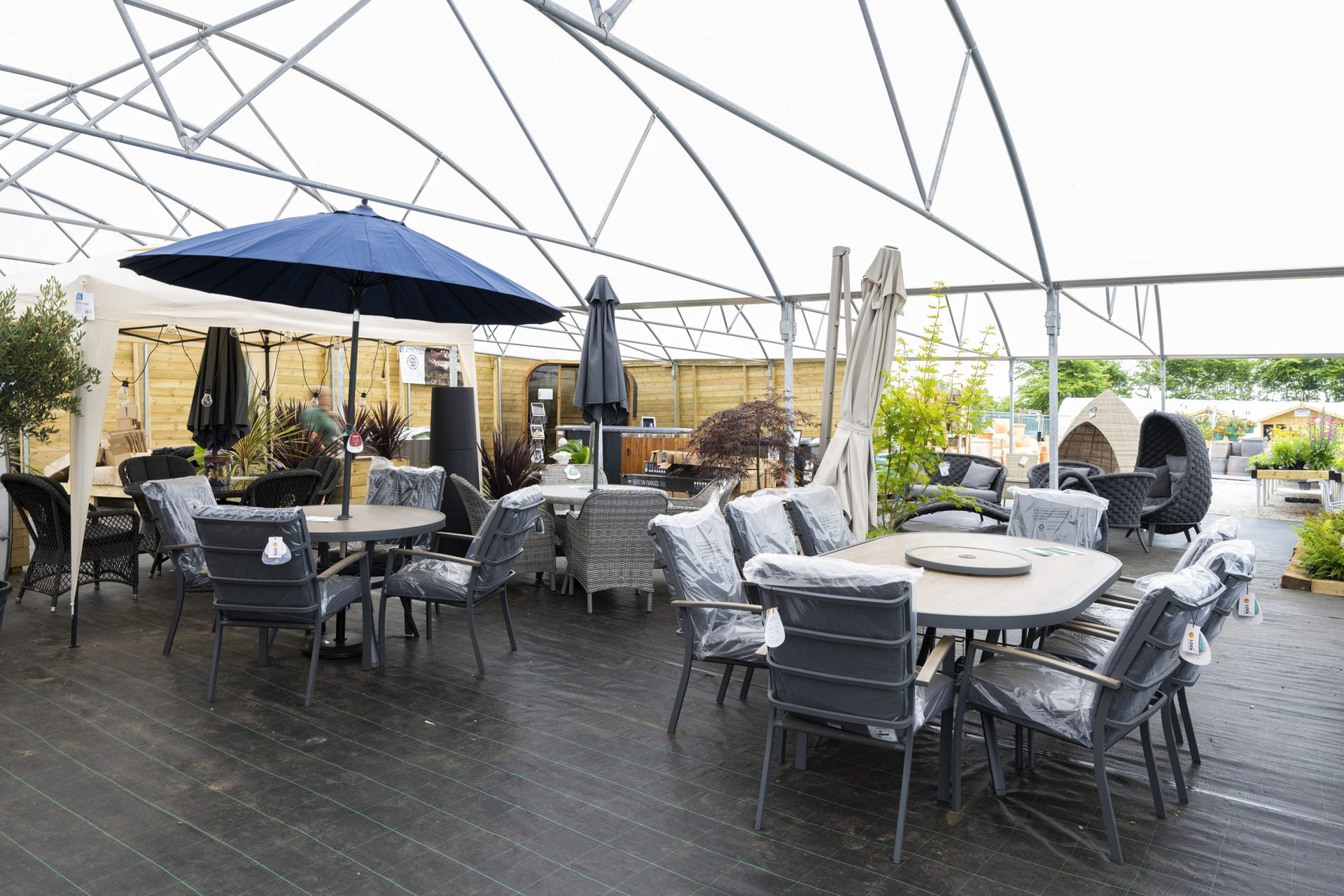 Outdoor furniture display under a white canopy; tables, chairs, umbrellas, with green plants in the background.