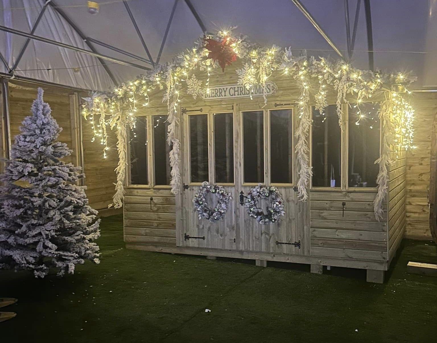 A festive wooden cabin decorated with lights, wreaths, and a snowy Christmas tree.