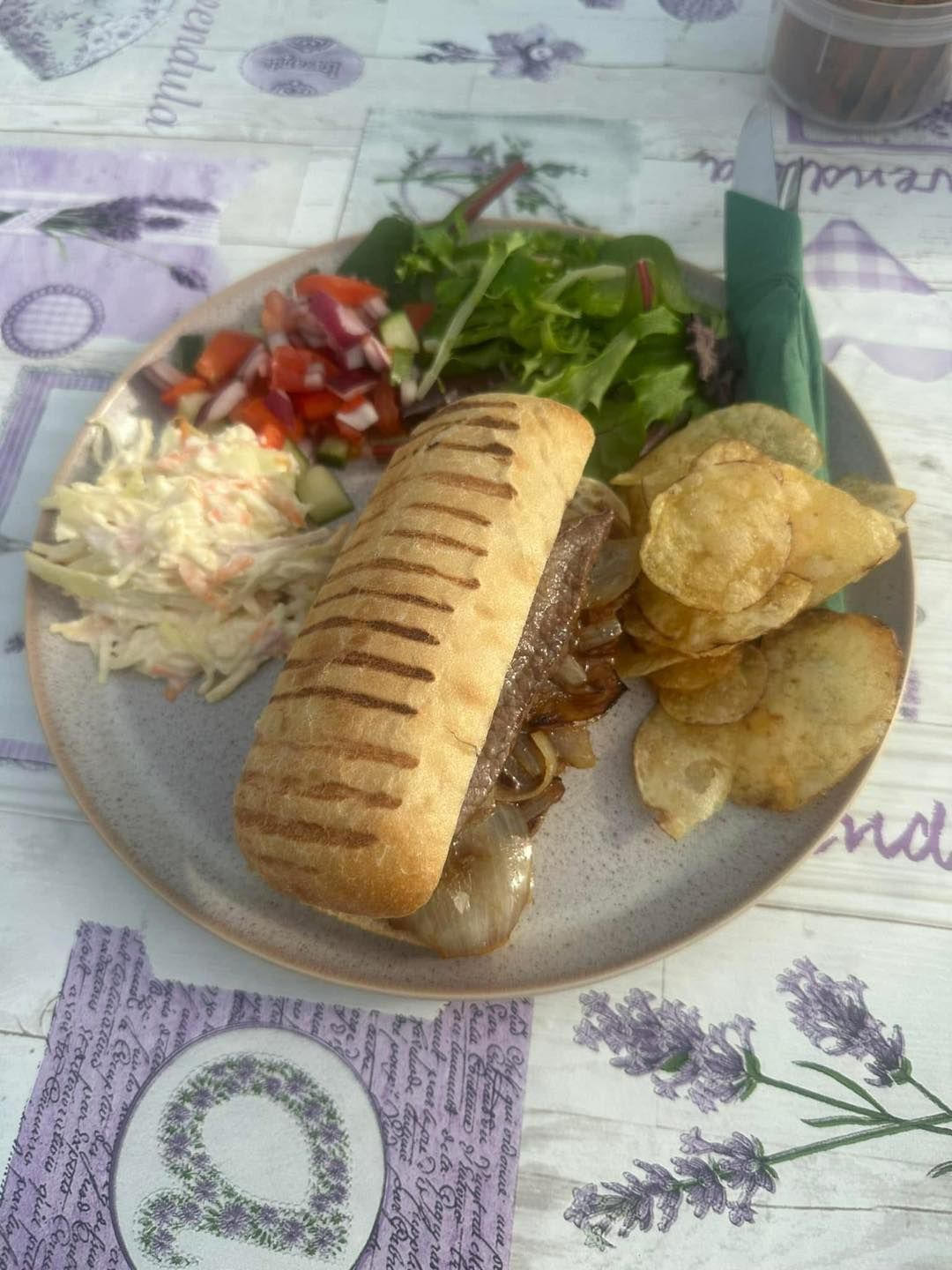 Sandwich with chips, salad, and coleslaw on a plate atop a lavender-patterned surface.