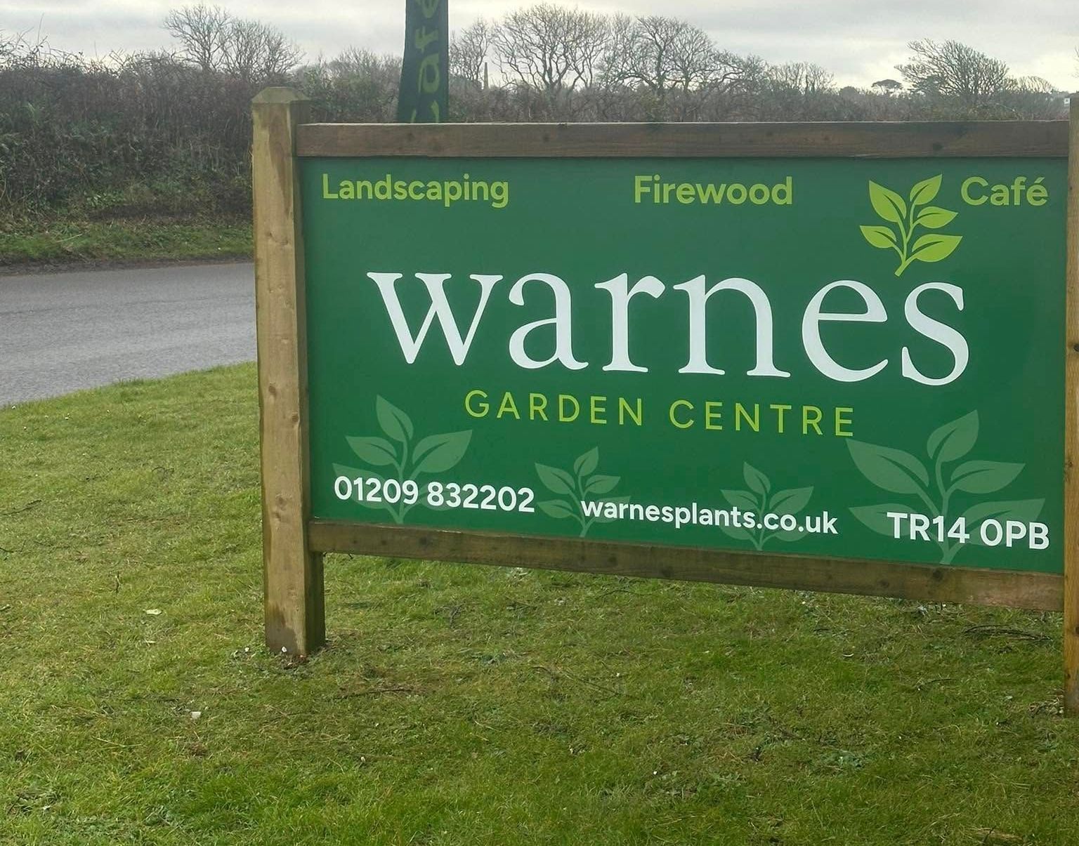 Sign for "Warnes Garden Centre" in green and white, on a green lawn; offers landscaping, firewood, and cafe.