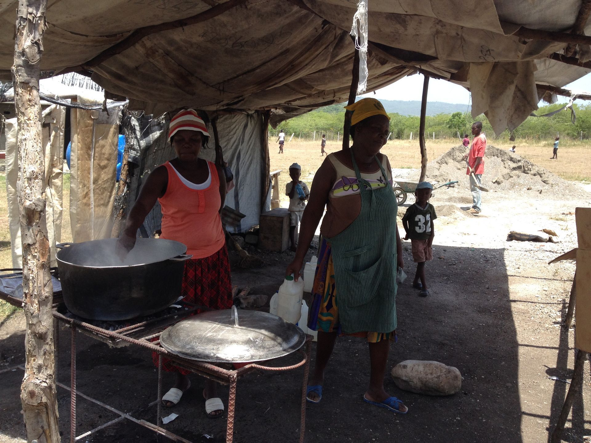 Two women cooking under a makeshift shelter; one stirs a pot, the other carries a jug.