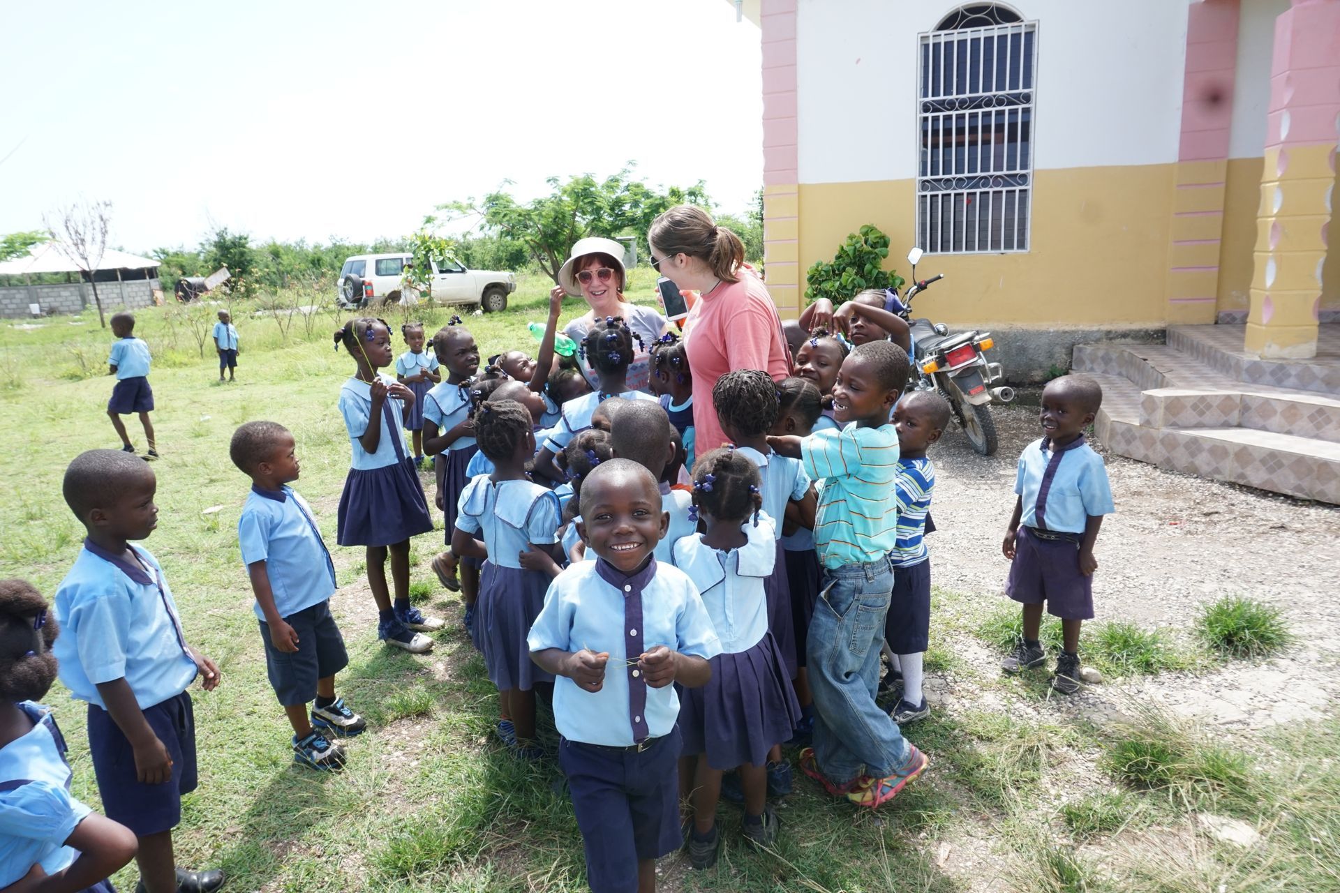 Children in blue uniforms gather around two women outside a building.