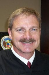 A man with a mustache is wearing a judge 's robe and smiling.