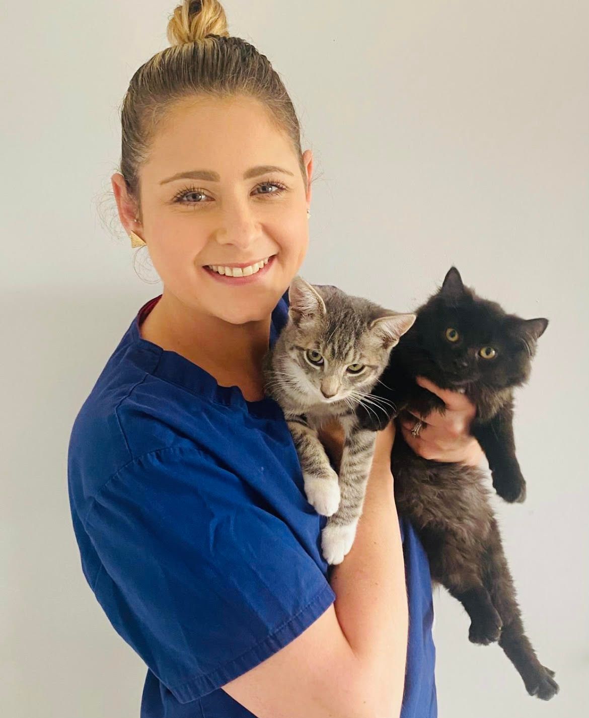 A woman in a blue scrub top is holding two cats in her arms.