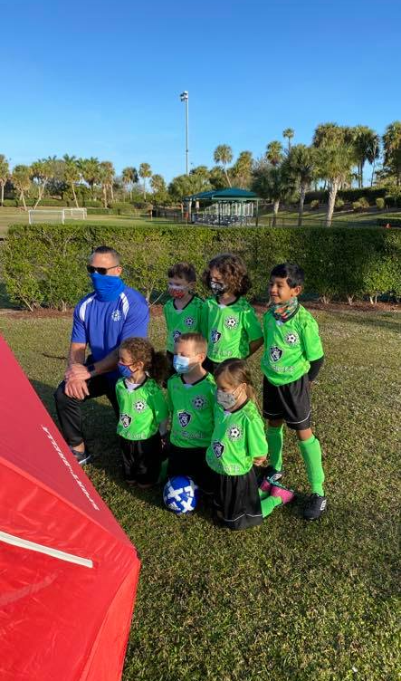 A group of young children wearing face masks are kneeling on a soccer field.