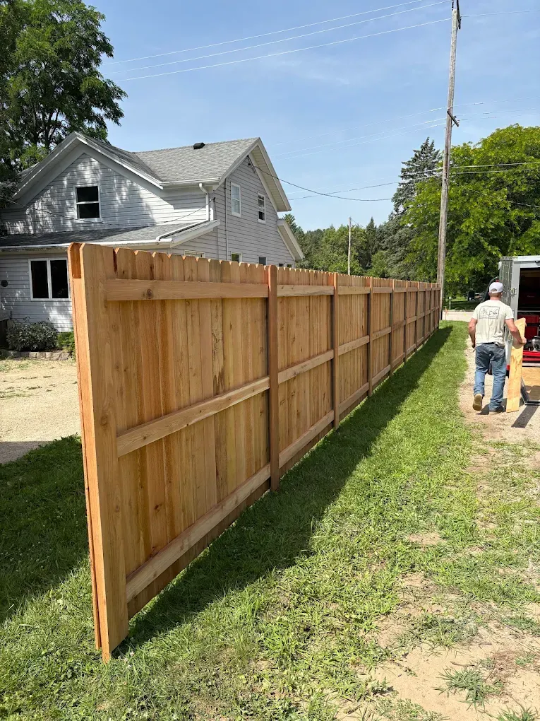 A wooden fence next to a grassy area with a house in the background and a person walking.