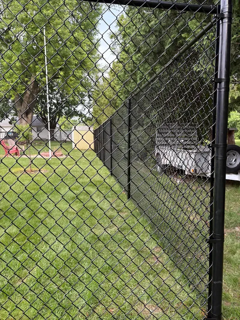 Black chain-link fence in green grass, with trees and a white trailer visible beyond.