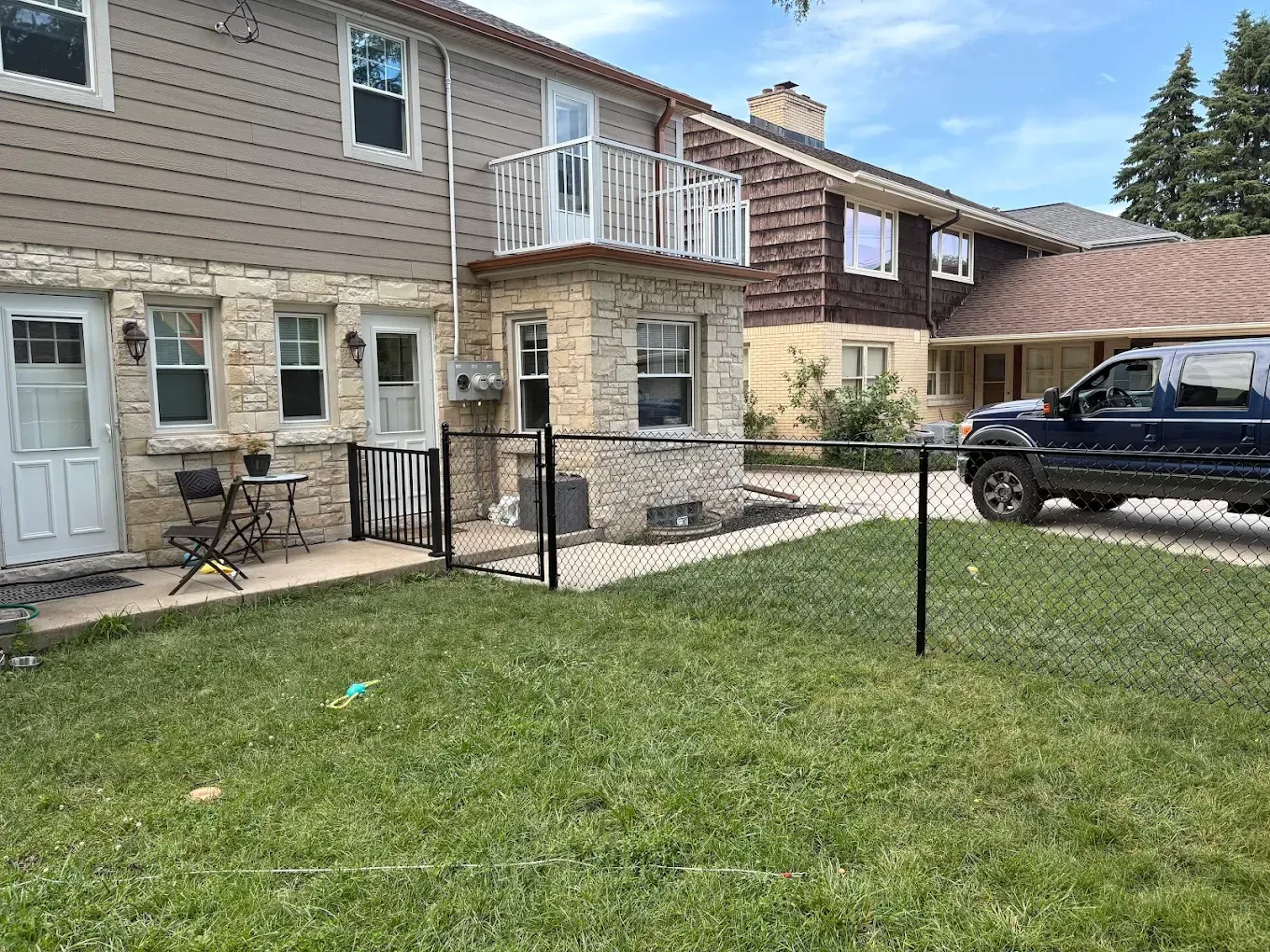 Exterior of a two-story beige house with a stone facade, a chain link fence in front, and a parked SUV.