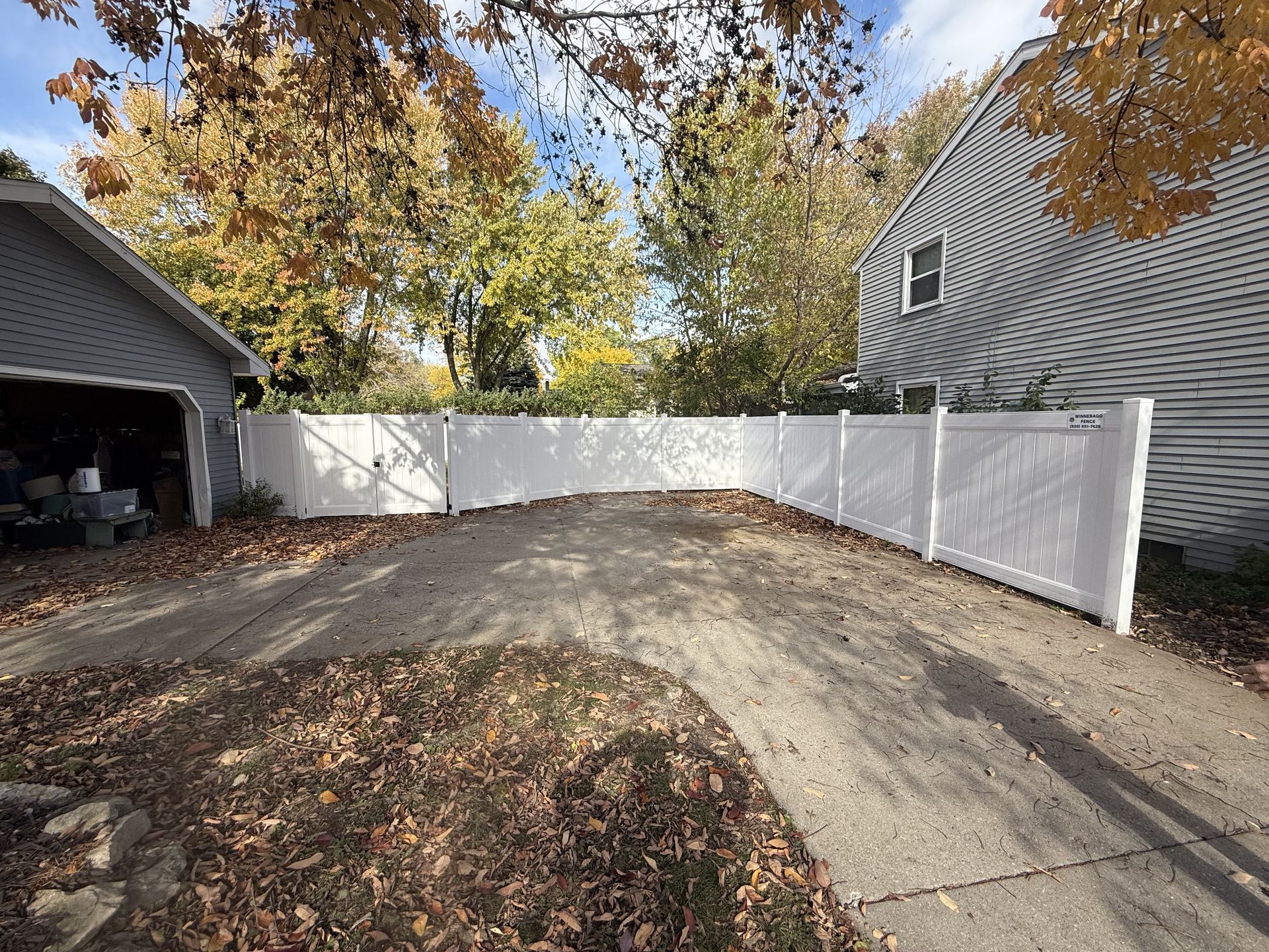 White vinyl fence enclosing a driveway, connecting to a garage and house. Fall foliage in the background.
