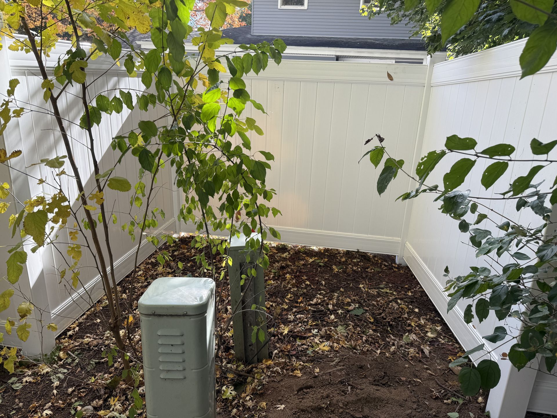 A fenced-in yard with utility boxes, trees, and brown mulch. White fence surrounds the area.