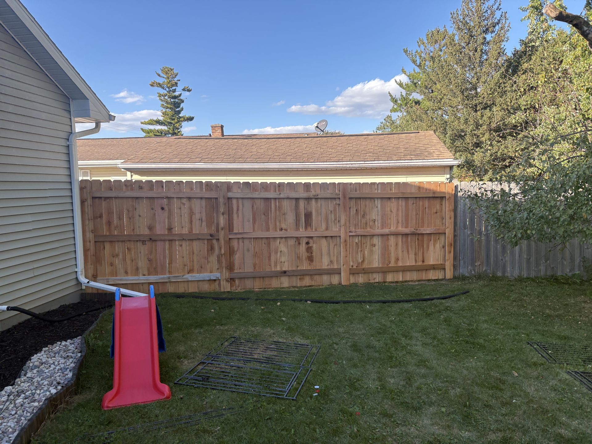 Backyard with red slide, brown wooden fence, green grass, and a clear blue sky.
