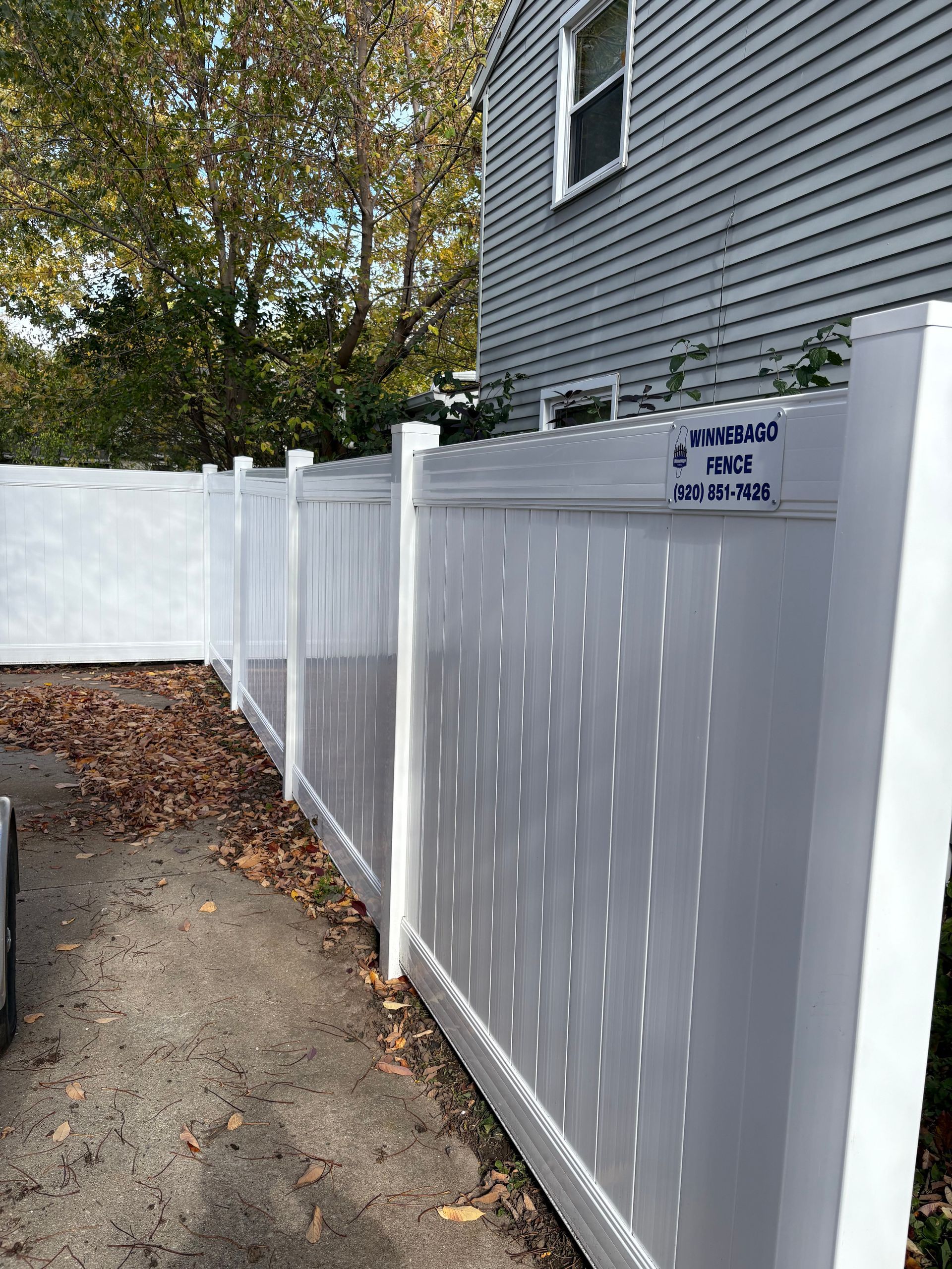 White vinyl fence along a driveway next to a gray house with a window and a label.