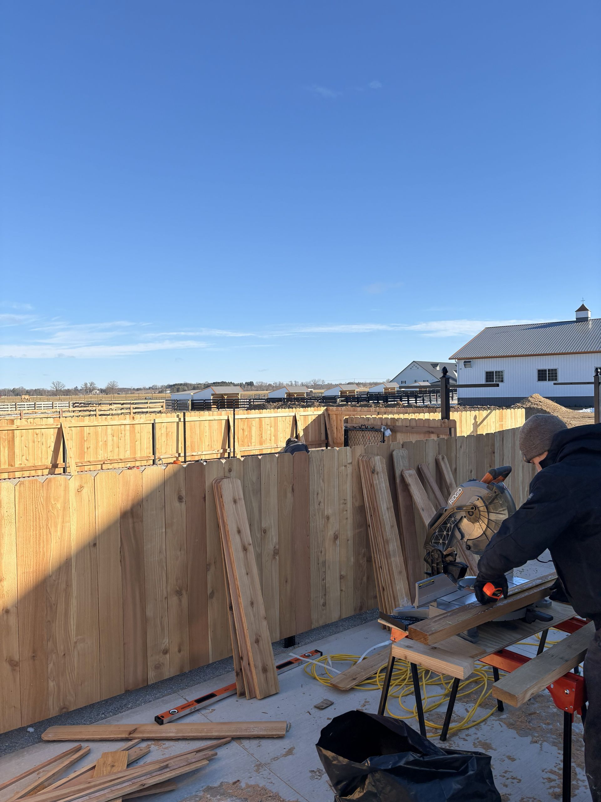 Person cutting wood with a saw; wooden fence under construction in daylight with a clear blue sky.