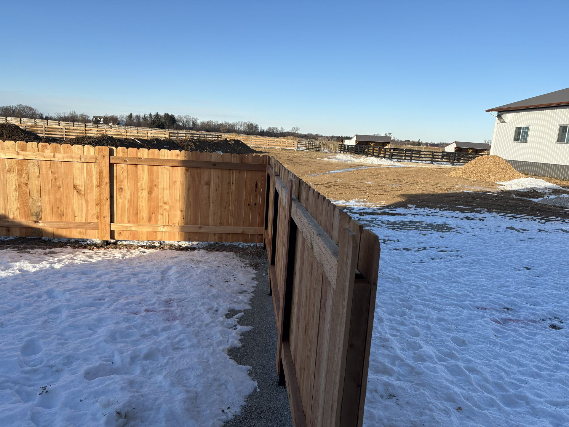 Wooden fence with snow-covered ground in front. Open field with dirt and a house in the background. Bright blue sky.