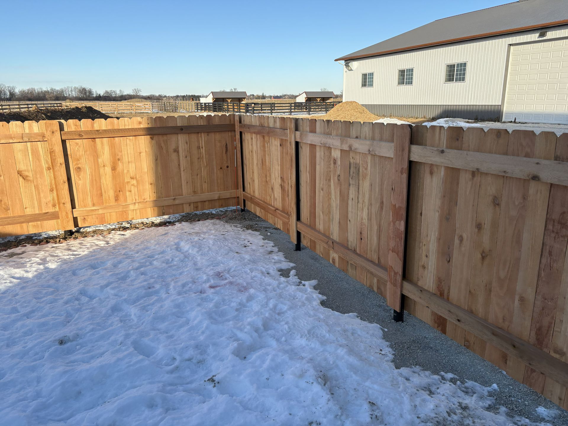 Wooden fence surrounding snow-covered yard near a house on a sunny day.