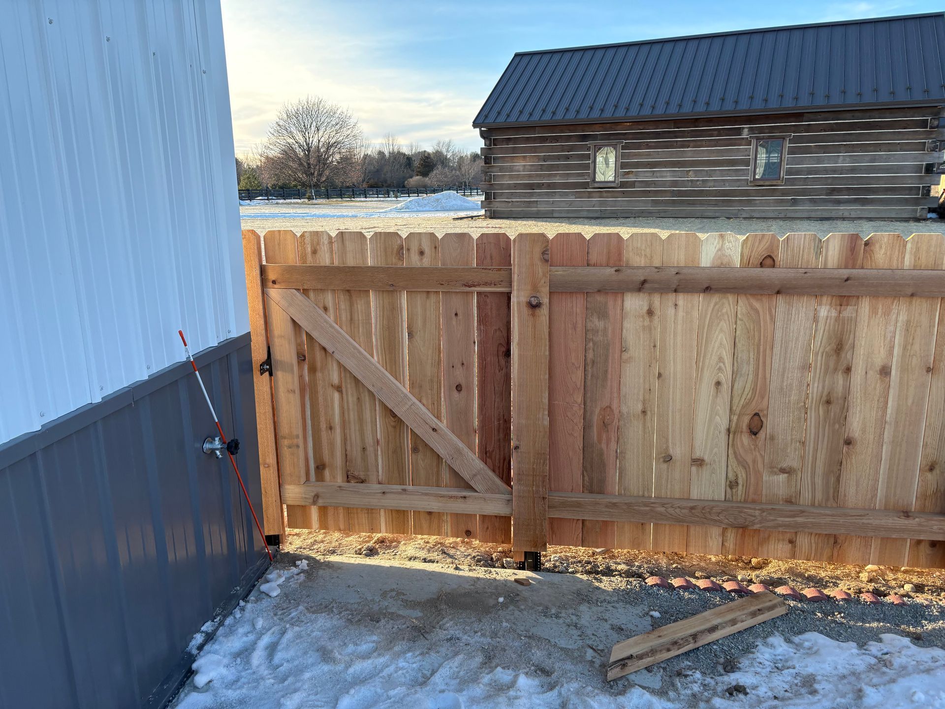 Wooden fence with gate, snow on the ground, adjacent to a gray building and a log cabin.