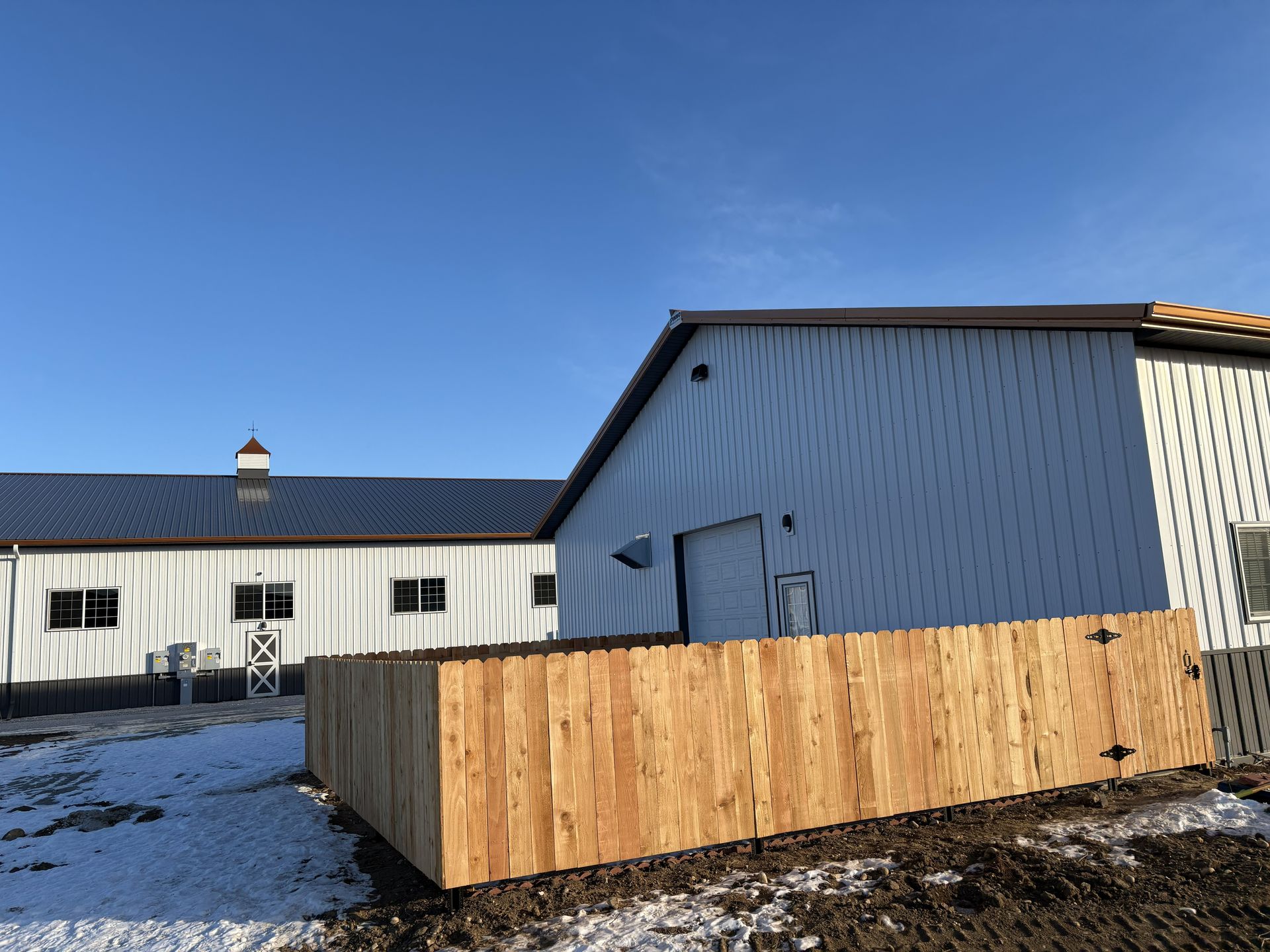 Two white barns with dark roofs, one with a wooden fence in front, against a blue sky.