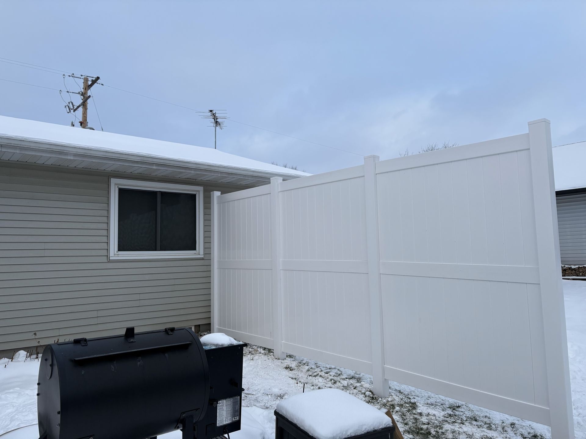 Backyard scene with snow, featuring a white fence, a house, and a black grill. Overcast sky.