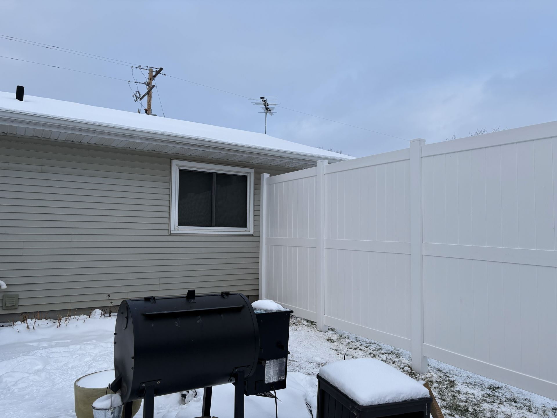 Backyard with snow, black grill, white fence, and a house under a cloudy sky.