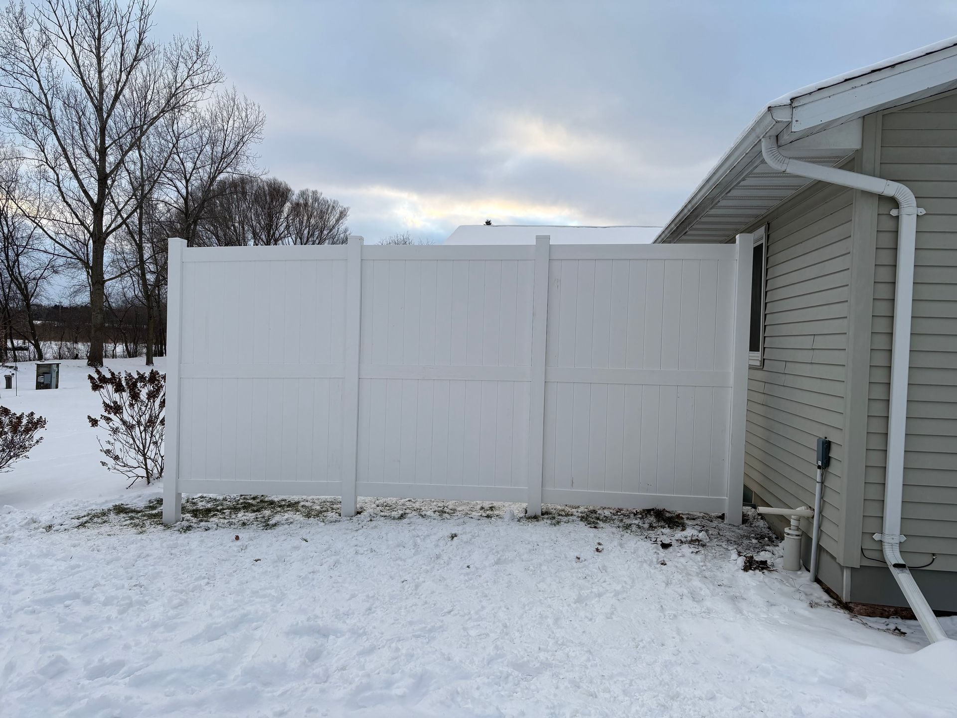 White vinyl fence next to a house with snow on the ground and a cloudy sky.