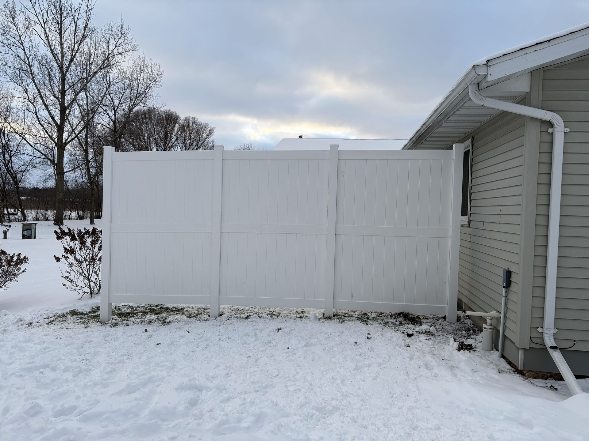 White vinyl fence next to a house with snow on the ground; cloudy sky.