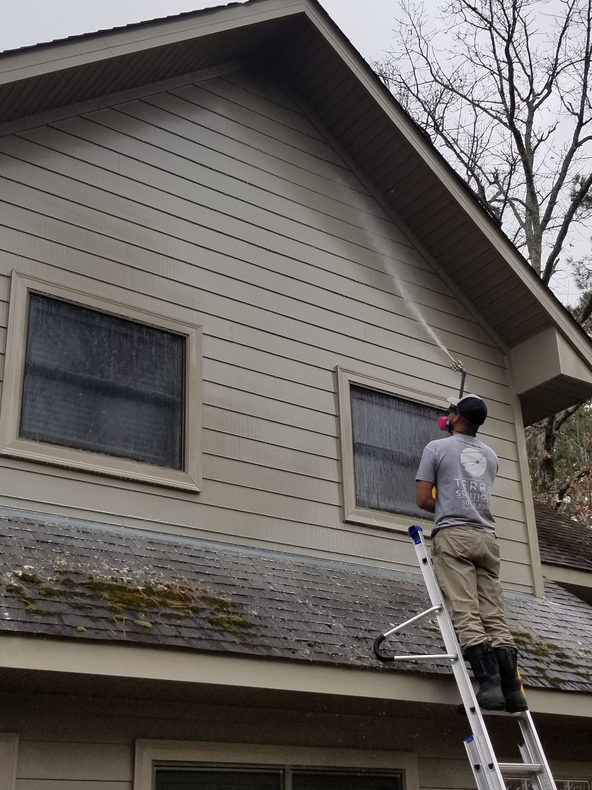 A man is standing on a ladder spraying water on the side of a house.