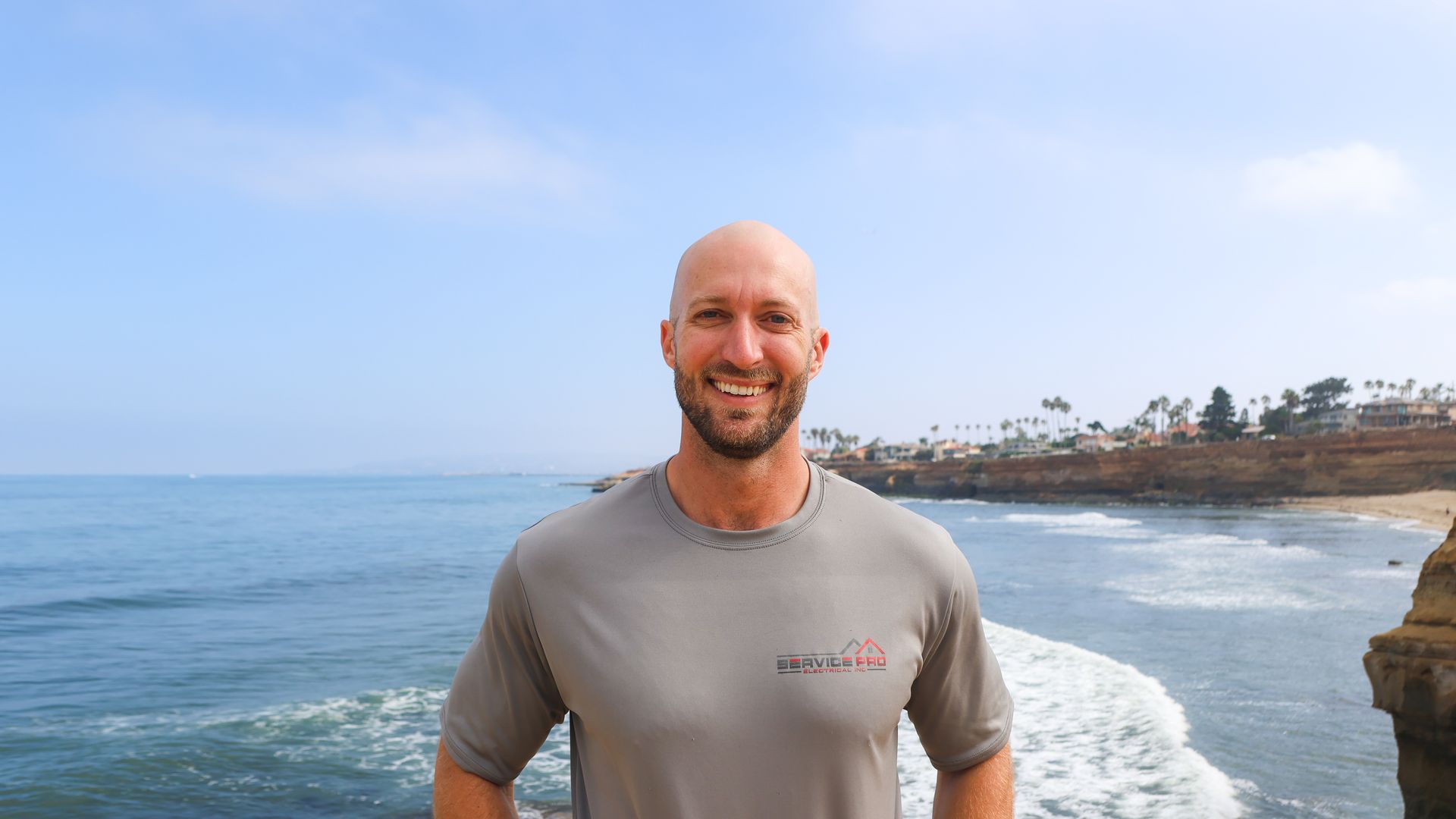 A bald man with a beard is standing in front of the ocean.