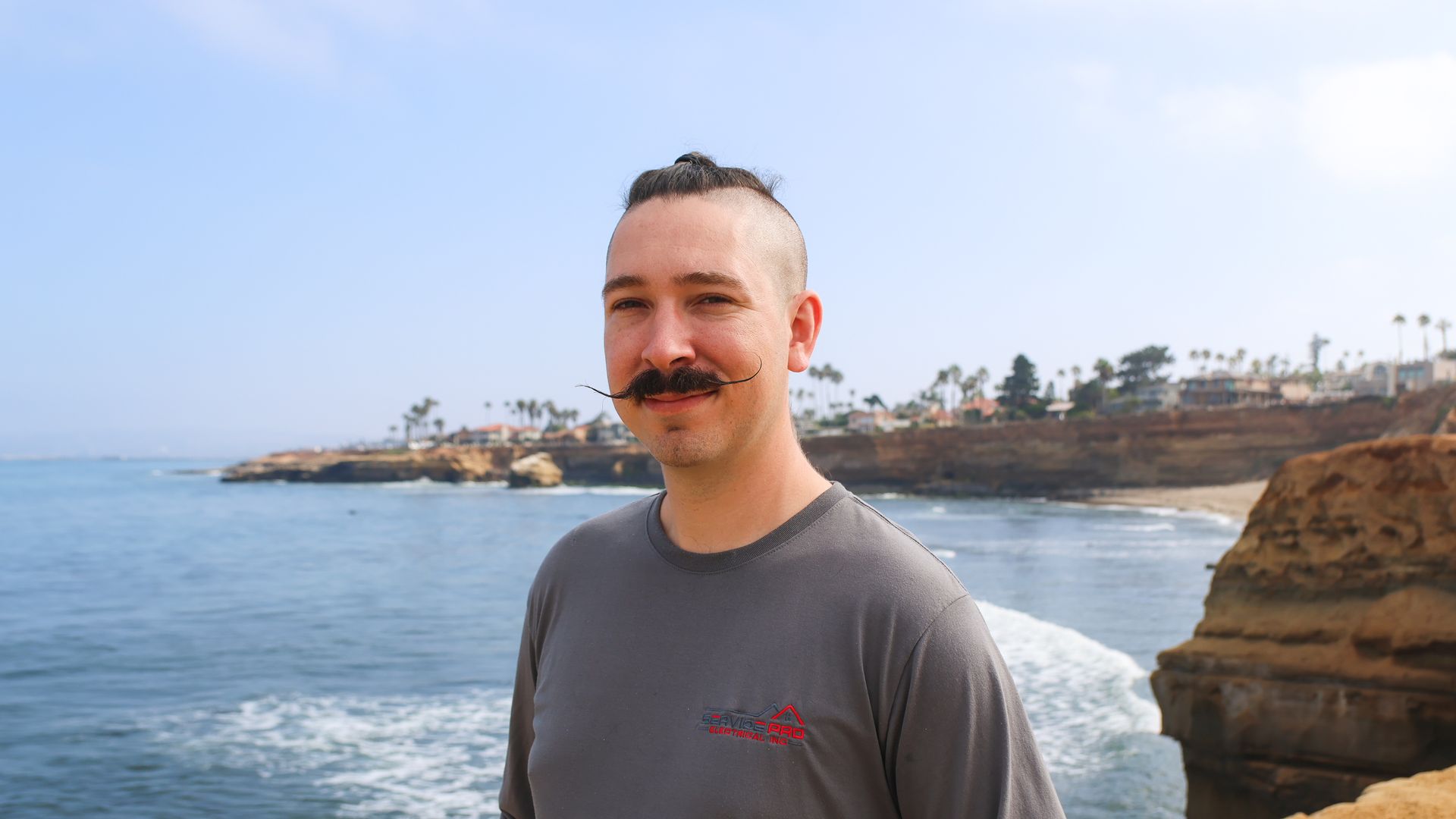 A man with a mustache is standing on a cliff overlooking the ocean.