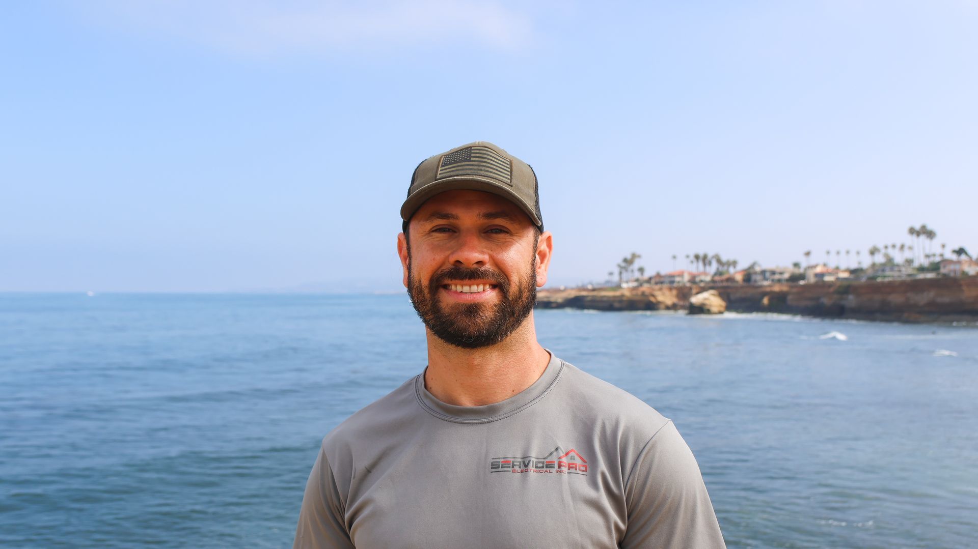 A man with a beard is standing in front of the ocean.