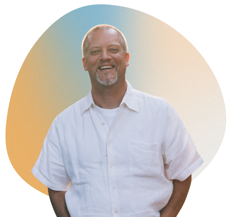 Man smiling, hands in pockets, wearing white shirt, soft background with pastel colors.