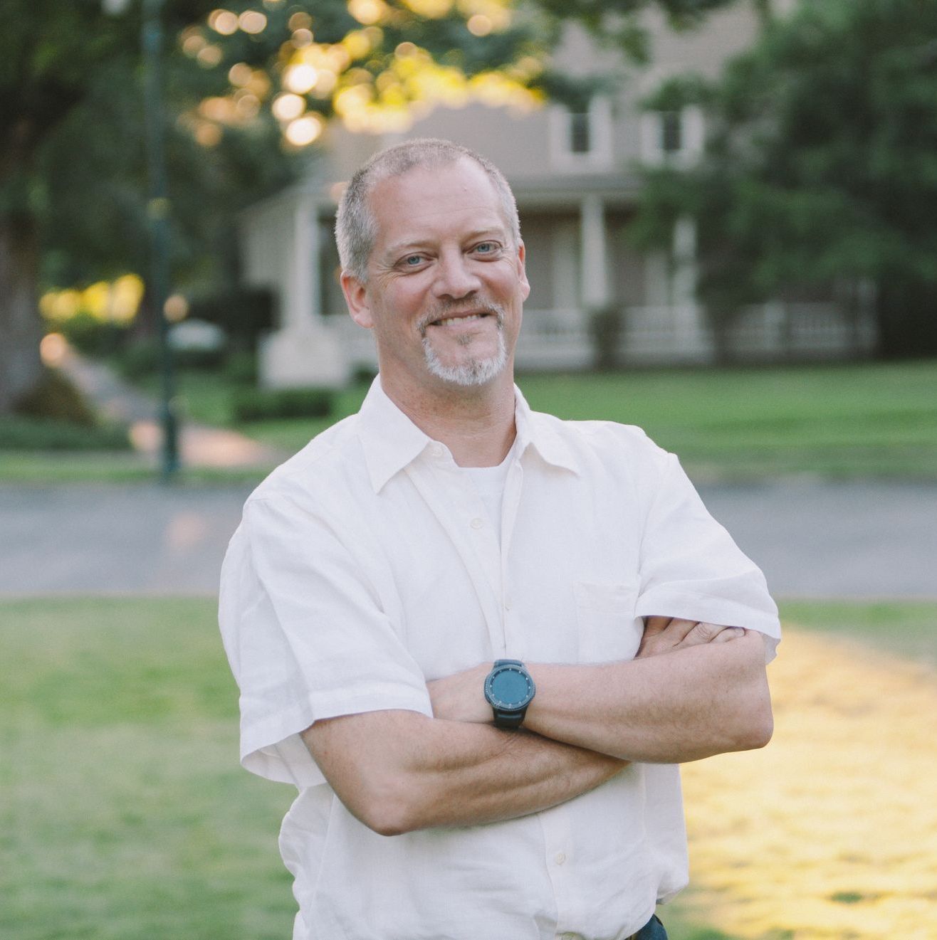 Man with crossed arms, smiling, in front of a house on a sunny day.