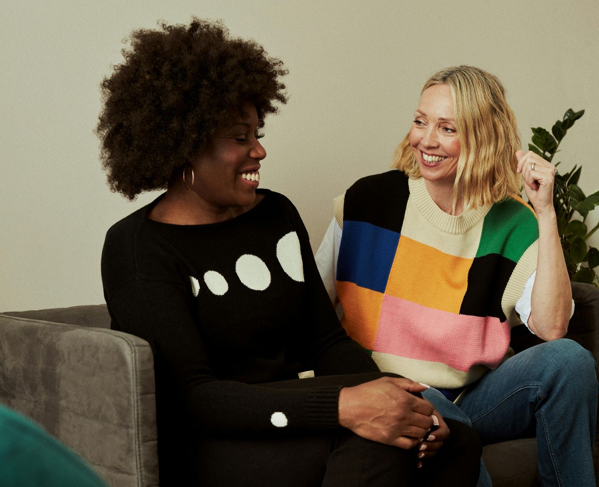 Two smiling women in a room; one with dark hair in a black sweater, the other with blonde hair in a colorful patchwork sweater.